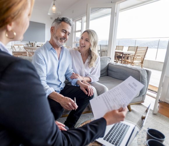 Happy mature couple meeting investments and financial advisor at home. They are happy and smiling sitting in the living room. The advisor is holding a document. There is a laptop on the table